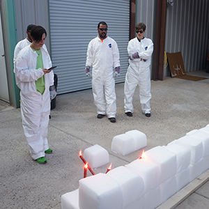 Color photograph of Jessica Silverman, David Linares, Rusty Johnson, and Megan Schulz standing in front of a row of white blocks with red flares stuck in them