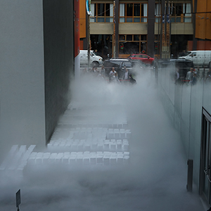 Color photograph of letters spelled in white blocks of dry ice emanating fog in a walkway between two buildings