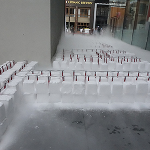 Color photograph of rows of white blocks of dry ice studded with flares and emanating fog in a walkway between two buildings