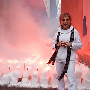 Color photograph of Judy Chicago standing in front of rows of white blocks of dry ice studded with red flares glowing and emanating fog