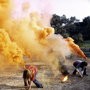 Color photograph of two people crouching surrounded by plumes of organe smoke