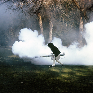 Color photograph of a figure carrying equipment emitting clouds of white smoke against a background of trees
