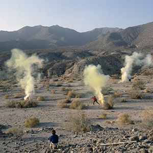 Color photograph of two figures amid rising plumes of yellow smoke in a desert landscape with hills in the background