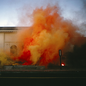 Color photograph of red and orange smoke rising in front of a beige building