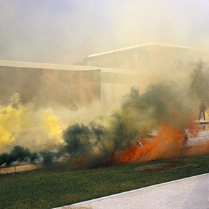 Color photograph of yellow, green, and orange smoke rising in front of a large building
