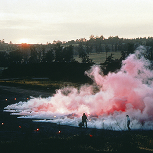 Color photograph of two figures standing next to pink and white smoke rising from a ring of flares on a grassy field