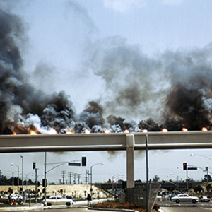 Color photograph of black smoke rising from flares on an overpass above a street