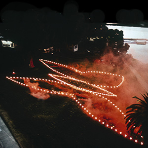 Color photograph of red smoke rising from flares in the shape of a butterfly at night