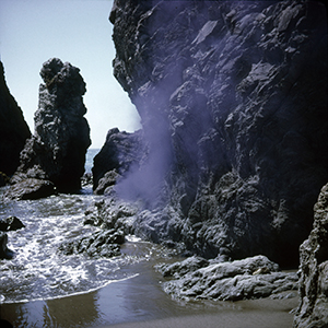Color photograph of purple smoke rising against a rocky cliff with water at the base