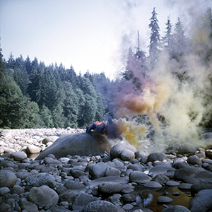 Color photograph of purple, pink, orange, and yellow smoke rising near a boulder in a rocky stream bed through a forest
