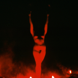 Color photograph of a nude woman raising her arms in the air bathed in red light with smoke at her feet against a black background