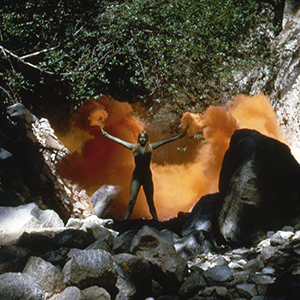 Color photograph of a nude woman standing with her arms and legs splayed, holding two flares from which orange smoke emerges in a rocky forest setting