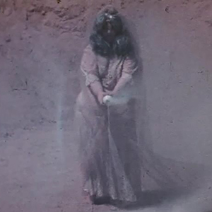 Color photograph of a woman wearing a beige, floor-length dress holding a white canister from which smoke emerges against a rocky background