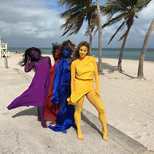 Color photograph of four women, each wearing a different color dress with their skin painted to match, standing together on a beach with palm trees in the background