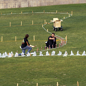 Color photograph of Chris Souza and another person kneeling surrounded by stakes and a curving white outline on a grassy field
