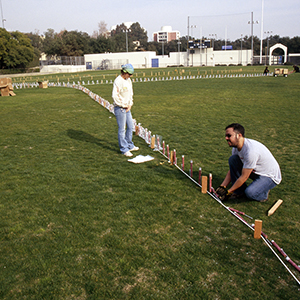 Color photograph of Rusty Johnson and another person kneeling and standing near a row of stakes and a white outline on a grassy field