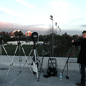 Color photograph of a person standing alongside five cameras on tripods on a white rooftop overlooking a football field