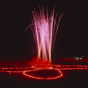 Color photograph of fireworks rising from red flares in the shape of a butterfly on a dark ground