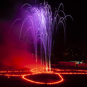 Color photograph of purple fireworks rising from red flares in the shape of a butterfly on a dark ground