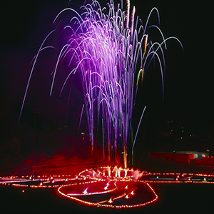 Color photograph of purple fireworks rising from red flares in the shape of a butterfly on a dark ground