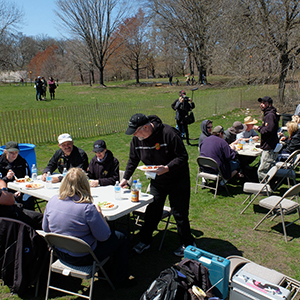 Color photograph of several groups of people eating at folding tables and chairs in a park