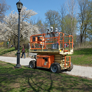 Color photograph of an orange mechanical lift parked next to a lamppost in a park