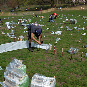 Color photograph of people setting up flares on a lawn strewn with items covered in plastic