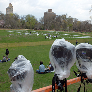Color photograph of three cameras wrapped in plastic overlooking a lawn with people working amid a white outline and scattered objects