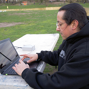 Color photograph of a person sitting and using a laptop on a table inside a fence on a lawn