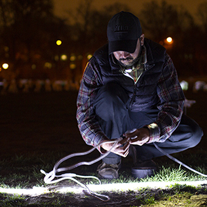 Color photograph of Rusty Johnson kneeling and holding wires on the grass at night