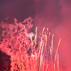 Color photograph of an aerial view of pink smoke and fireworks shooting up from a glowing red outline of a butterfly on a lawn in a park