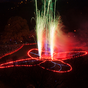 Color photograph of an aerial view of fireworks shooting up from a glowing red outline of a butterfly with blue interior lines on a lawn in a park as a crowd looks on