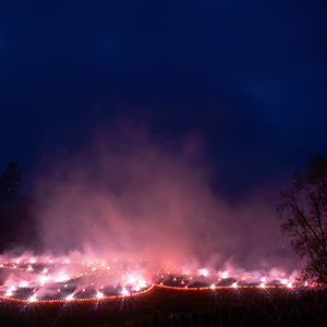 Color photograph of white smoke emanating from a glowing red butterfly outline studded with white lights in a darkened park