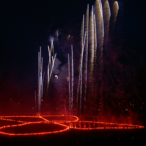 Color photograph of fireworks shooting up from a glowing red outline of a butterfly in a darkened park
