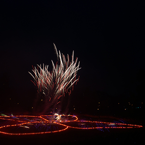 Color photograph of fireworks shooting up from a glowing red outline of a butterfly in a darkened park