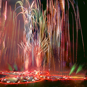 Color photograph of multi colored fireworks shooting up from a glowing red outline of a butterfly in a darkened park