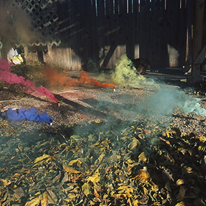 Color photograph of five differently colored plumes of smoke emanating from gravel with wooden structures in the background