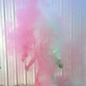 Color photograph of nude Katie Schroeder holding red and green smoke flares with similar flares planted in the ground at her feet