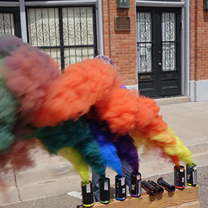 Color photograph of Judy Chicago standing behind multicolored smoke emerging from canisters on a wooden support in front of a brick building