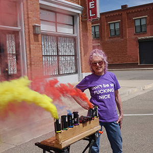 Color photograph of Judy Chicago next to multicolored smoke emerging from canisters on a wooden support in front of a brick building