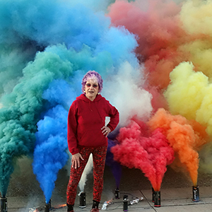 Color photograph of Judy Chicago standing in front of multicolored plumes of smoke