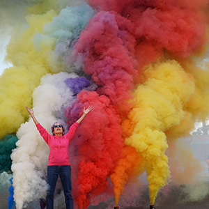Color photograph of Judy Chicago standing with her arms raised in front of multicolored plumes of smoke