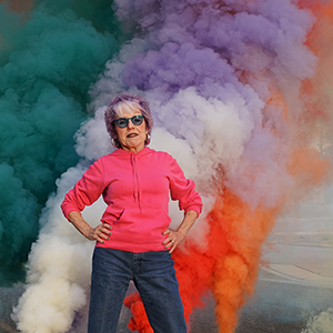 Color photograph of Judy Chicago standing with her hands on her hips in front of multicolored plumes of smoke