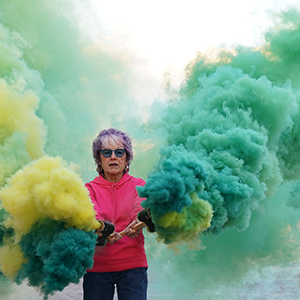 Color photograph of Judy Chicago holding canisters mounted on wood from which green and yellow smoke is emanating