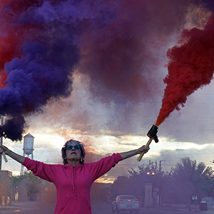 Color photograph of Judy Chicago standing on a city street with her arms raised holding two canisters mounted on wood from which purple and red smoke is emanating
