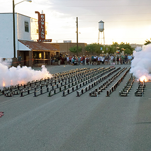 Color photograph of white smoke rising from rows of smoke canisters in the middle of a paved road as a crowd looks on at dusk