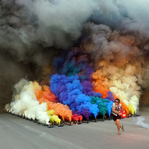 Color photograph of Mary Costa running beside plumes of multicolored smoke rising from rows of smoke canisters in the middle of a paved road