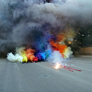 Color photograph of a strip of firecrackers exploding beside plumes of multicolored smoke rising from rows of smoke canisters in the middle of a paved road