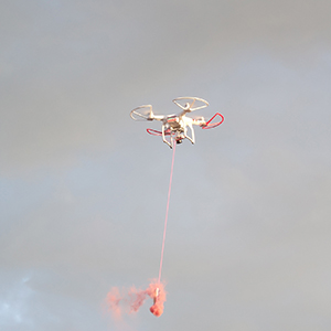 Color photograph of a drone carrying an orange smoke canister in a cloudy sky