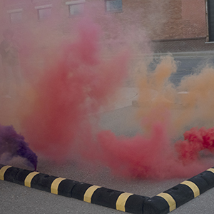 Color photograph of plumes of purple, red, and orange smoke rising within black and yellow barriers on a paved street